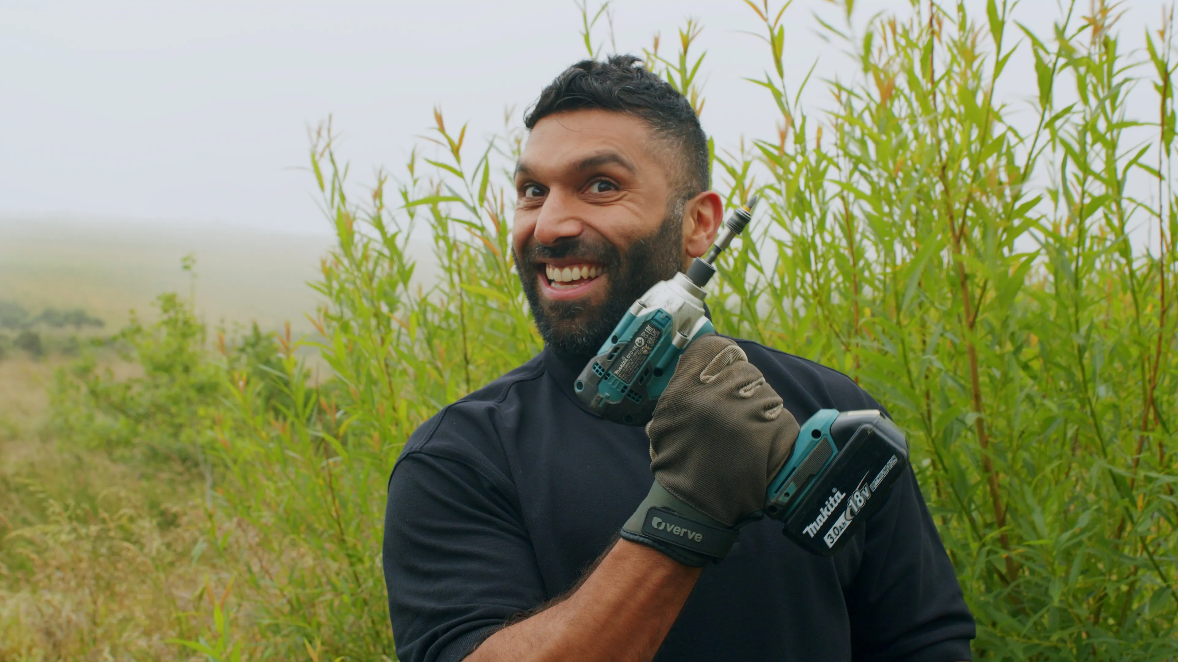 A smiling man in a black shirt and gloves holds a cordless drill in an outdoor setting with lush green plants, conveying enthusiasm and readiness.